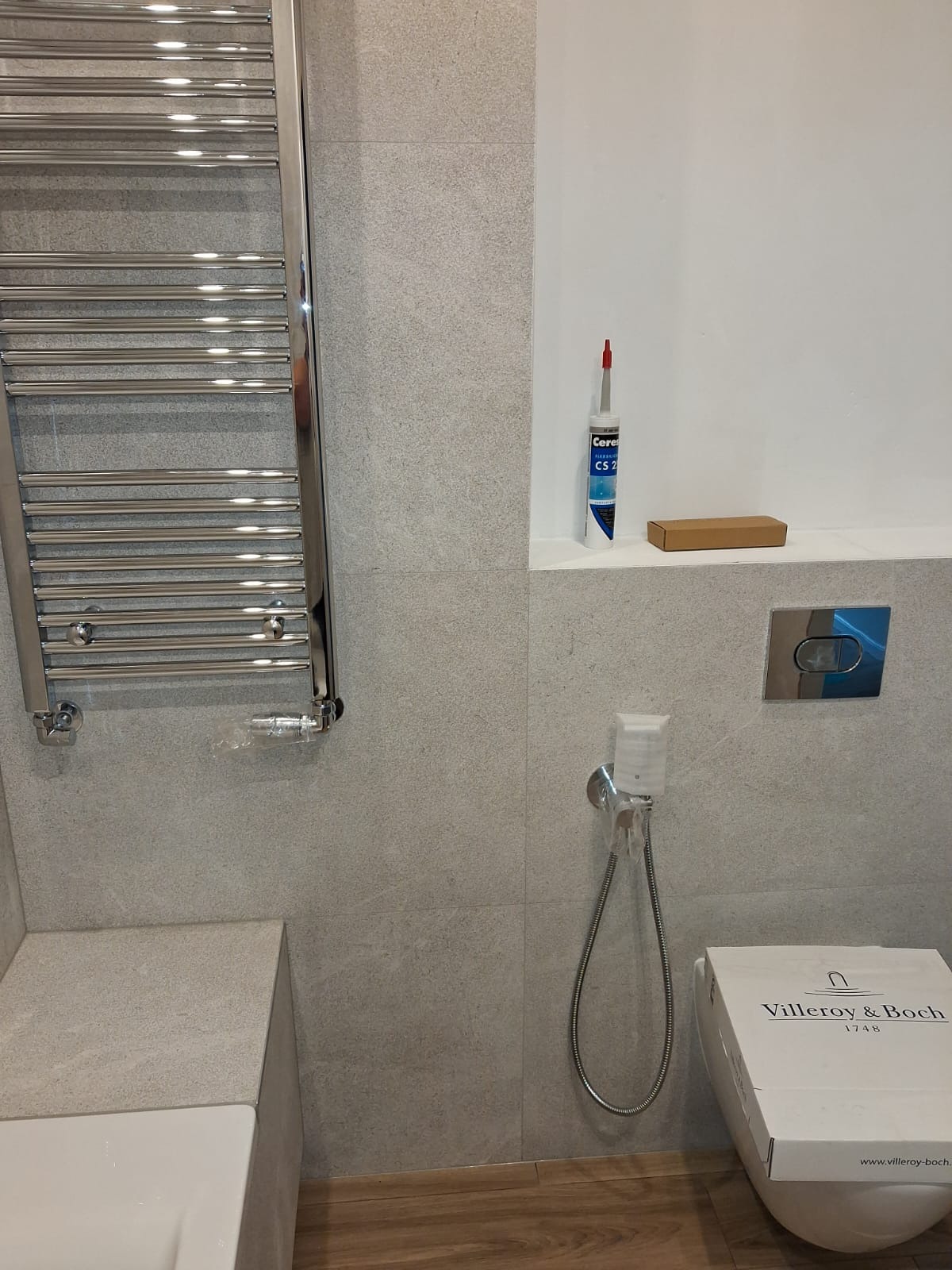 Wykończona łazienka z grzejnikiem drabinkowym, bidetką i toaletą Villeroy&Boch w kartonie. Na półce silikon Ceresit i kartonik. Płytki w odcieniach szarości.