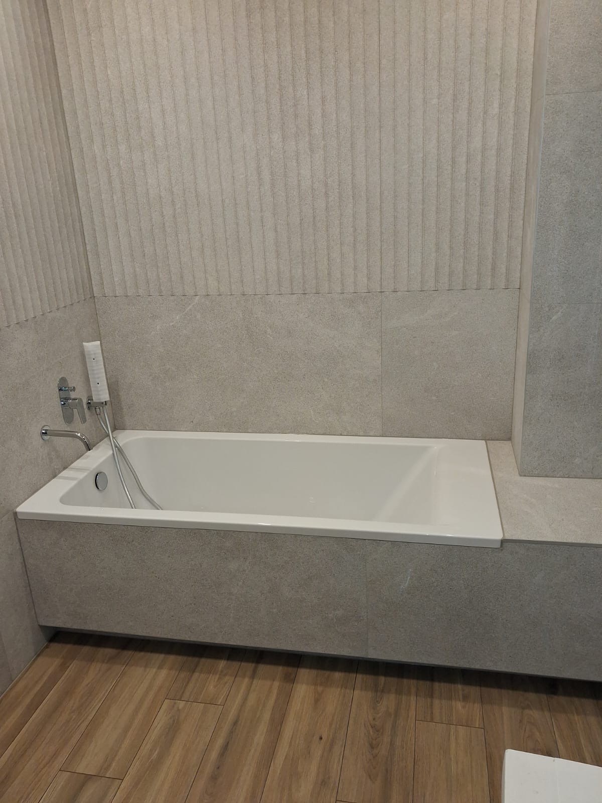 Nowoczesna, wykafelkowana wanna z hydromasażem i panelem prysznicowym w łazience. Beżowe płytki z delikatnym wzorem i drewnopodobna podłoga. Minimalistyczny design.