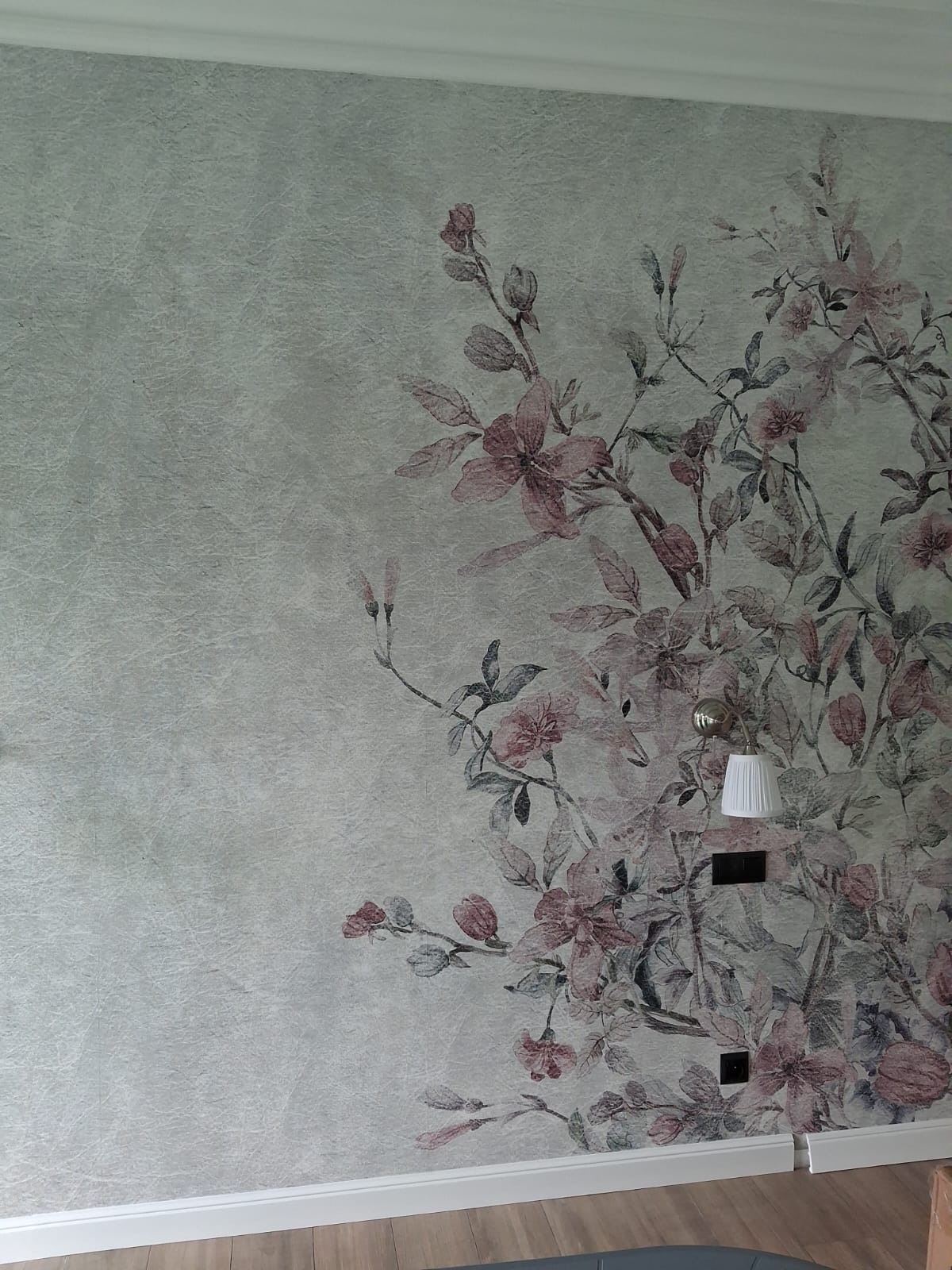 Ściana z dekoracyjną tapetą w kwiaty, oświetlona lampą ścienną. Widoczne gniazdka elektryczne i listwa przypodłogowa. Delikatna faktura tapety i stonowana kolorystyka.