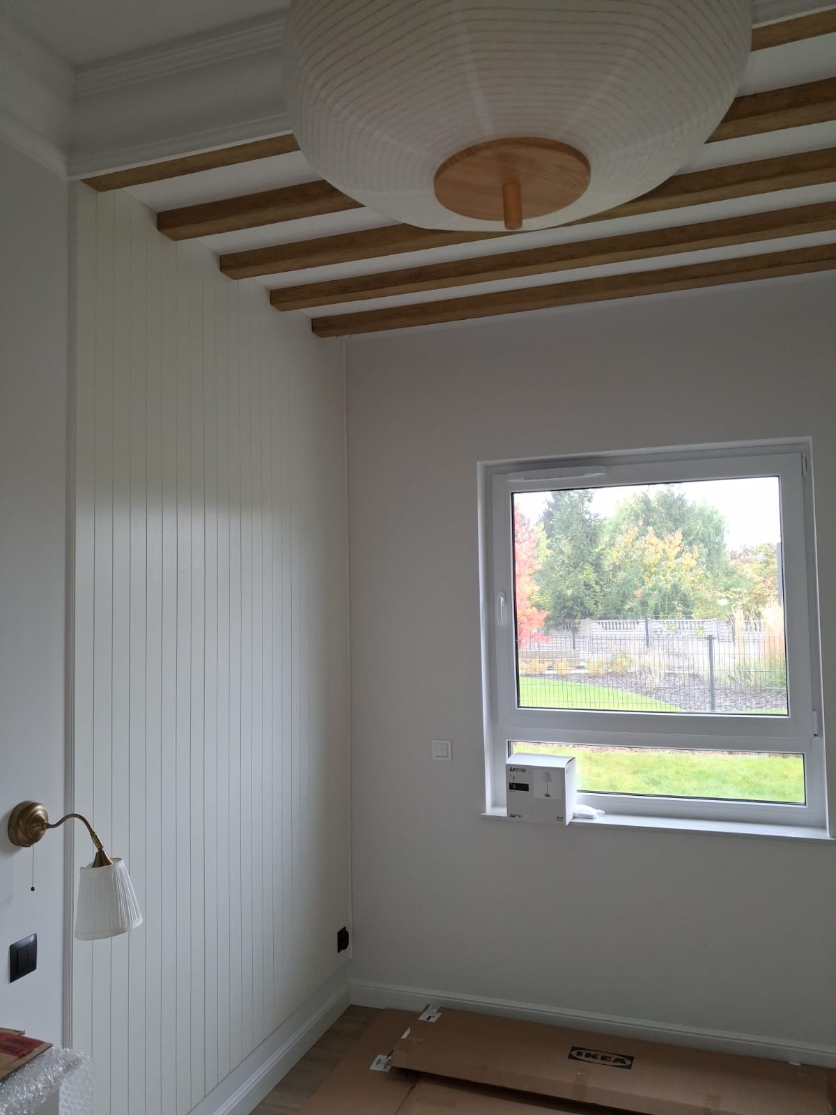 Białe panele ścienne z drewnianymi belkami sufitowymi i lampą. Widok na okno z zielenią. Kartony na podłodze sugerują etap wykończenia wnętrza.