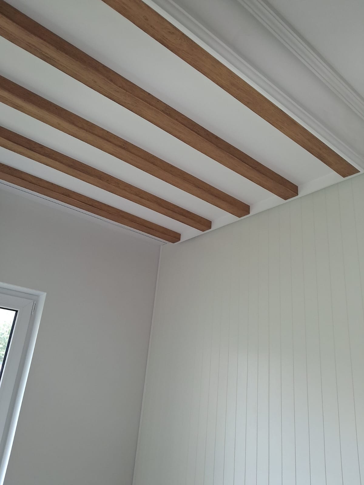 Biały sufit z dekoracyjnymi, drewnianymi belkami i listwami przypodłogowymi, w rogu widoczne okno i ściana z pionowymi panelami. Jasne wnętrze.