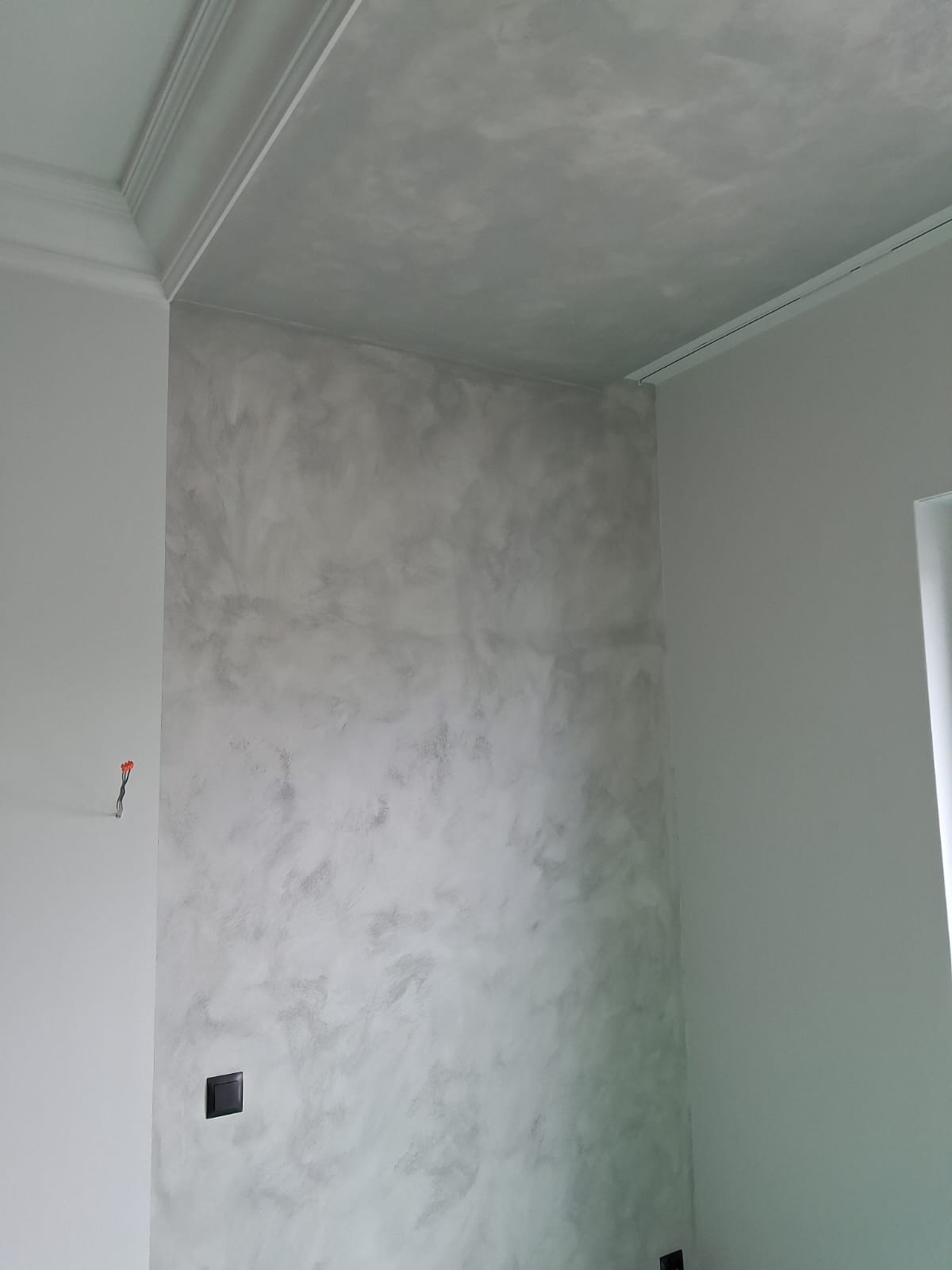 Ściana z efektem dekoracyjnym w odcieniach szarości, widoczna listwa sufitowa, gniazdko elektryczne i fragment okna. Minimalistyczne wykończenie wnętrza.