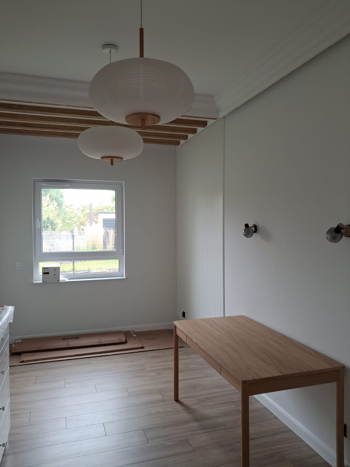 Jasne wnętrze pokoju z drewnianą podłogą i stołem. Widoczne lampy wiszące, okno i dekoracyjne listwy sufitowe. Kartony na podłodze sugerują etap wykończenia.