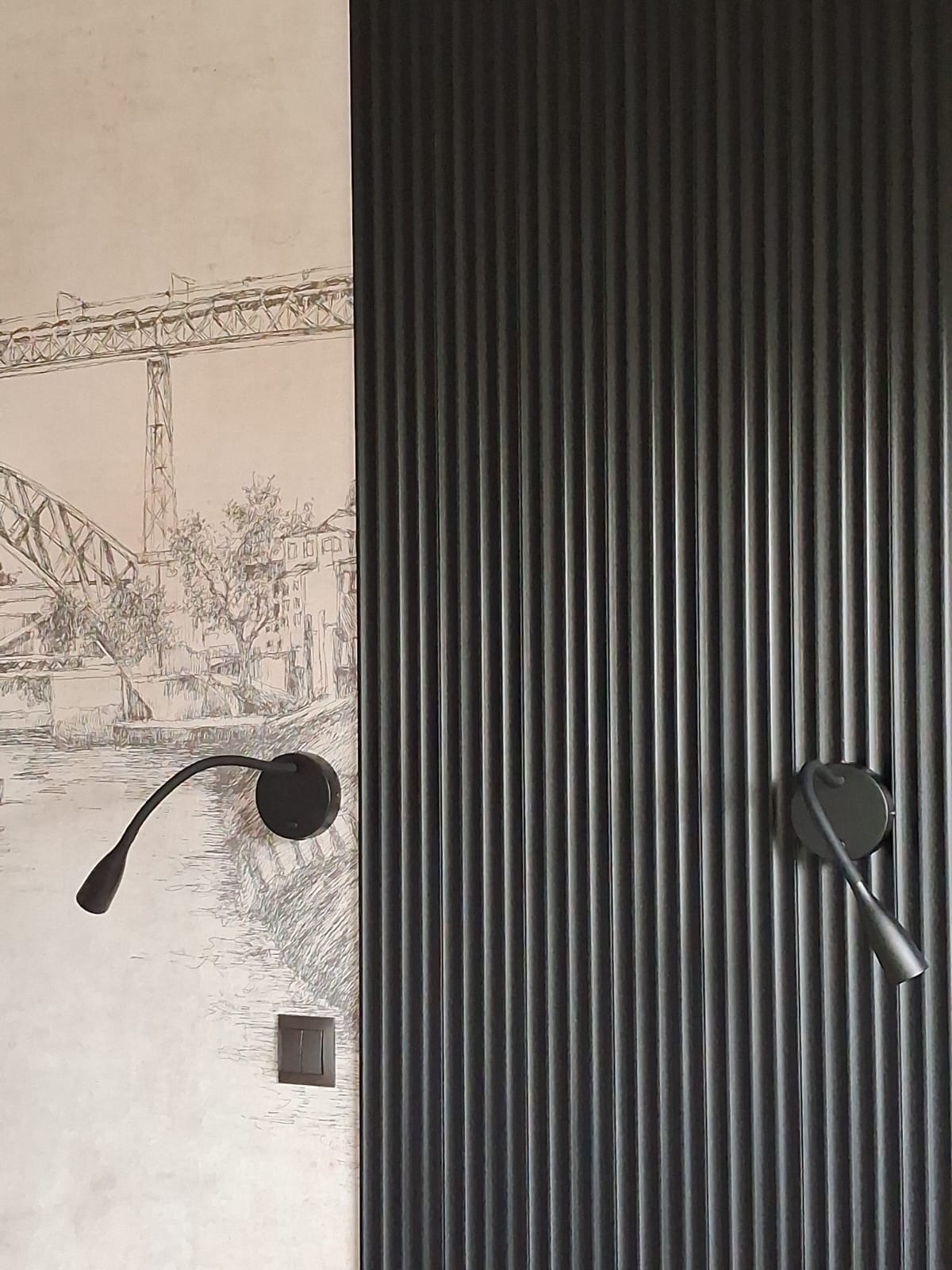 Nowoczesna ściana z czarnymi, pionowymi panelami dekoracyjnymi i lampką do czytania na tle ściany z rysunkiem mostu. Minimalistyczny design wnętrza.