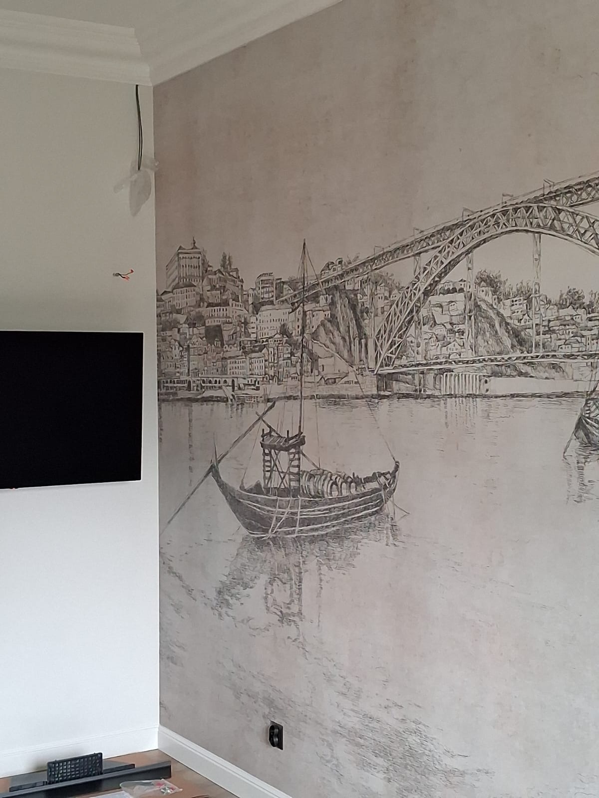 Ściana z fototapetą przedstawiającą krajobraz z łodzią i mostem, obok telewizor. Widoczne kable i listwa przypodłogowa. Szaro-biała kolorystyka.