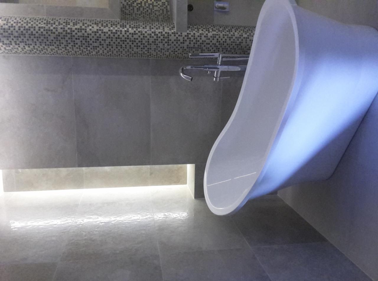 Nowoczesna łazienka z wolnostojącą wanną o asymetrycznym kształcie, szare płytki wielkoformatowe na ścianach i podłodze, dekoracyjna mozaika nad baterią umywalkową, podświetlenie LED pod szafką.