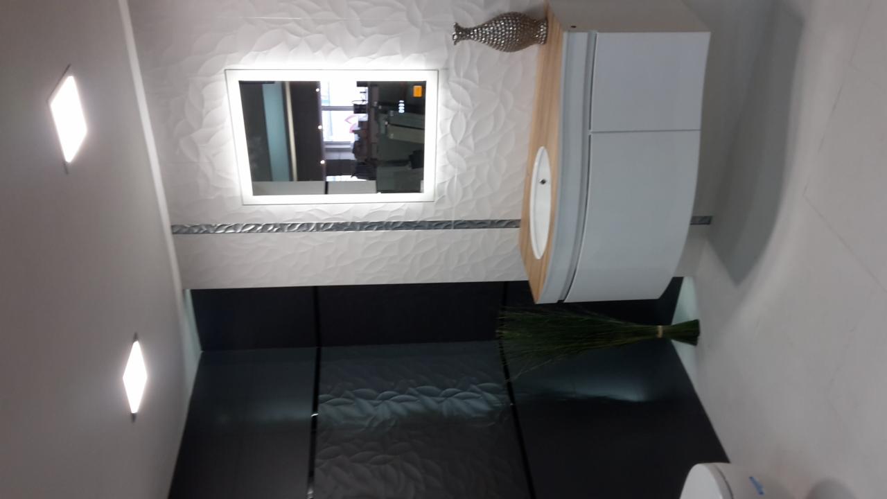 Nowoczesna łazienka z białymi, trójwymiarowymi płytkami na ścianie, podświetlanym lustrem, szafką z umywalką o drewnianym blacie i czarnymi płytkami podłogowymi z subtelnym wzorem.