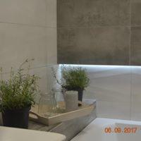 Minimalistyczna łazienka: fragment ściany z szarymi płytkami, podświetlona półka z drewnianą tacą, na której stoją dwie doniczki z ziołami, szklany słoik i biały kubek.