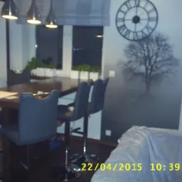 Nowoczesna jadalnia z ciemnym stołem, szarymi krzesłami barowymi, lampami wiszącymi i fototapetą z motywem drzewa oraz zegarem na ścianie.