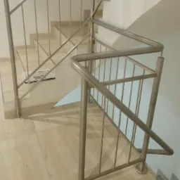 Metalowa konstrukcja zabezpieczająca schody wewnątrz budynku, z okrągłymi profilami i pionowymi prętami, widoczne płytki podłogowe i gazeta na schodach.