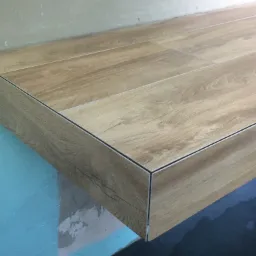 Wykończona półka z płytek imitujących drewno, z precyzyjnymi łączeniami i widoczną strukturą drewna, na tle surowej ściany w odcieniach błękitu i beżu.