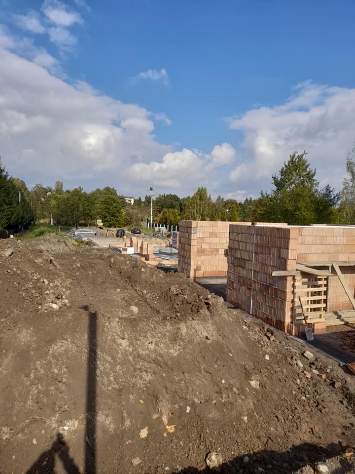 Budowa domu jednorodzinnego w trakcie wznoszenia ścian z czerwonej cegły, widoczny plac budowy z hałdą ziemi, w tle drzewa i błękitne niebo z chmurami.