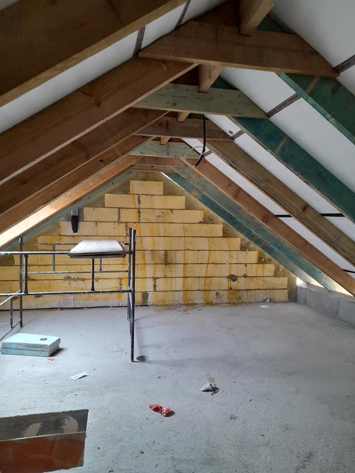 Surowe poddasze w trakcie adaptacji w Krakowie: widoczne drewniane krokwie, ocieplenie z żółtej wełny mineralnej ułożone schodkowo, fragment metalowej konstrukcji i betonowa posadzka.