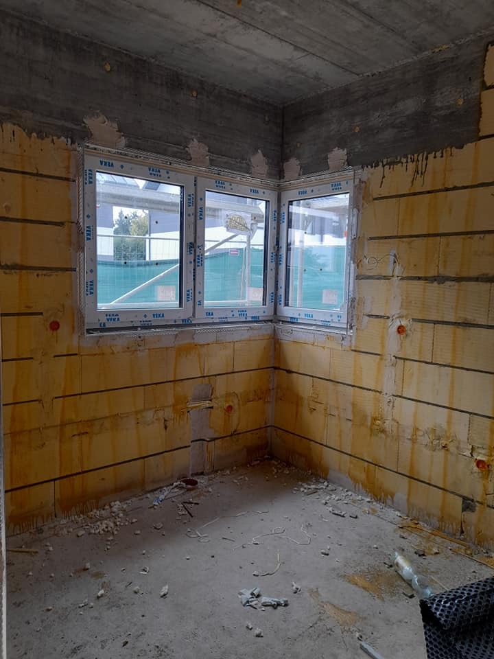 Surowy stan wnętrza z trzema oknami PCV marki VEKA w narożniku, ściany z izolacją z widocznymi poziomymi liniami, betonowy sufit, podłoga w stanie deweloperskim.