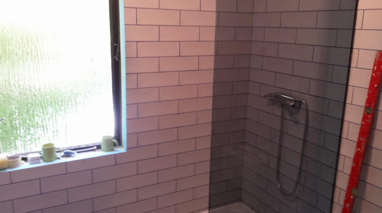 Świeżo wykafelkowana ściana łazienki z widocznym oknem, na parapecie dwie zielone filiżanki, poziomica oparta o ścianę kabiny prysznicowej, chromowana bateria prysznicowa.