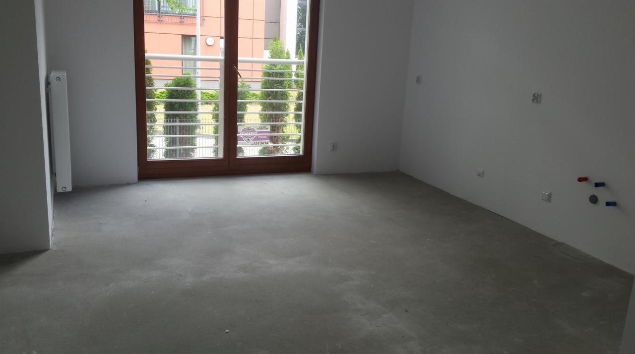 Surowe wnętrze pokoju przed wykończeniem: wylewka na podłodze, białe ściany z wystającymi instalacjami elektrycznymi i wodnymi, okno balkonowe z widokiem na zieleń.