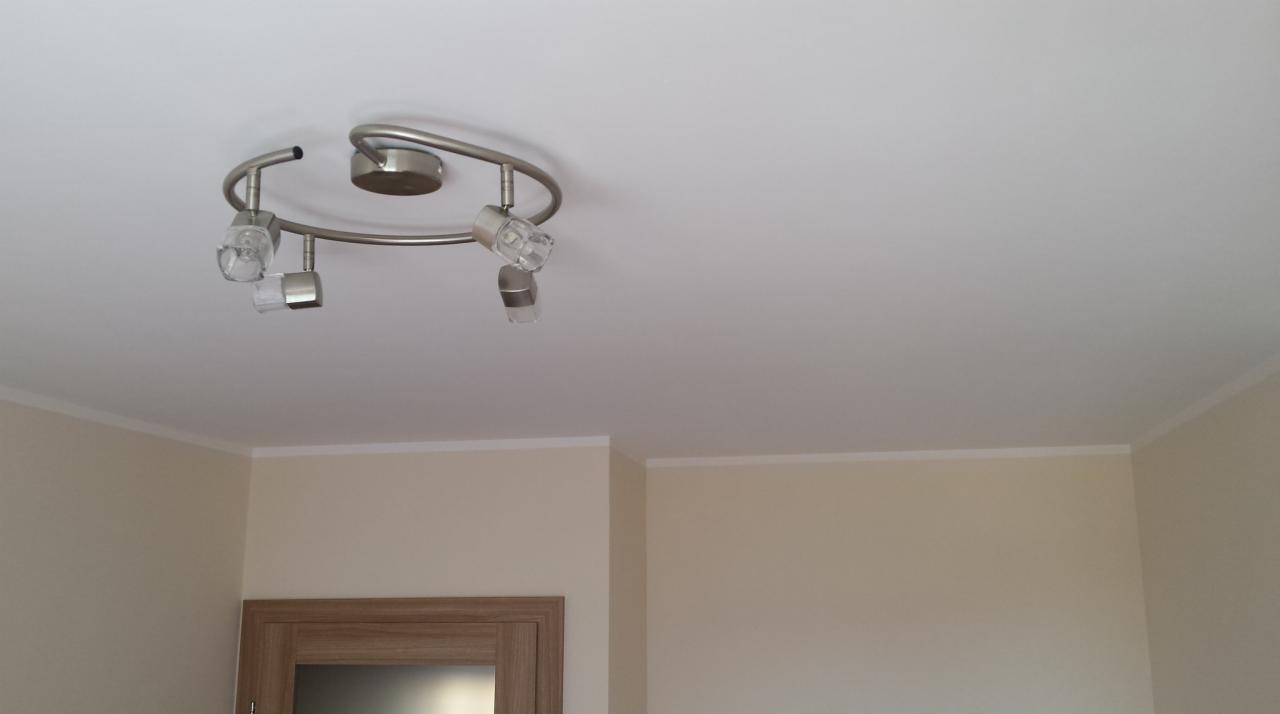 Srebrna lampa sufitowa z trzema reflektorami z przezroczystego szkła na tle białego sufitu i kremowych ścian z białą listwą przypodłogową.