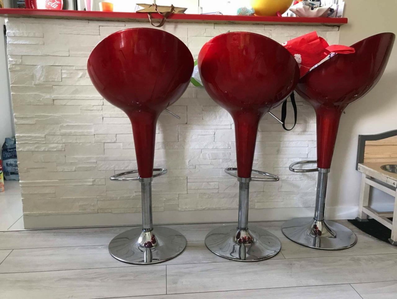 Trzy czerwone, błyszczące hokery barowe na chromowanych nogach stoją przed ścianą wyłożoną białymi płytkami imitującymi kamień. Widoczny fragment blatu barowego z czerwonym wykończeniem.