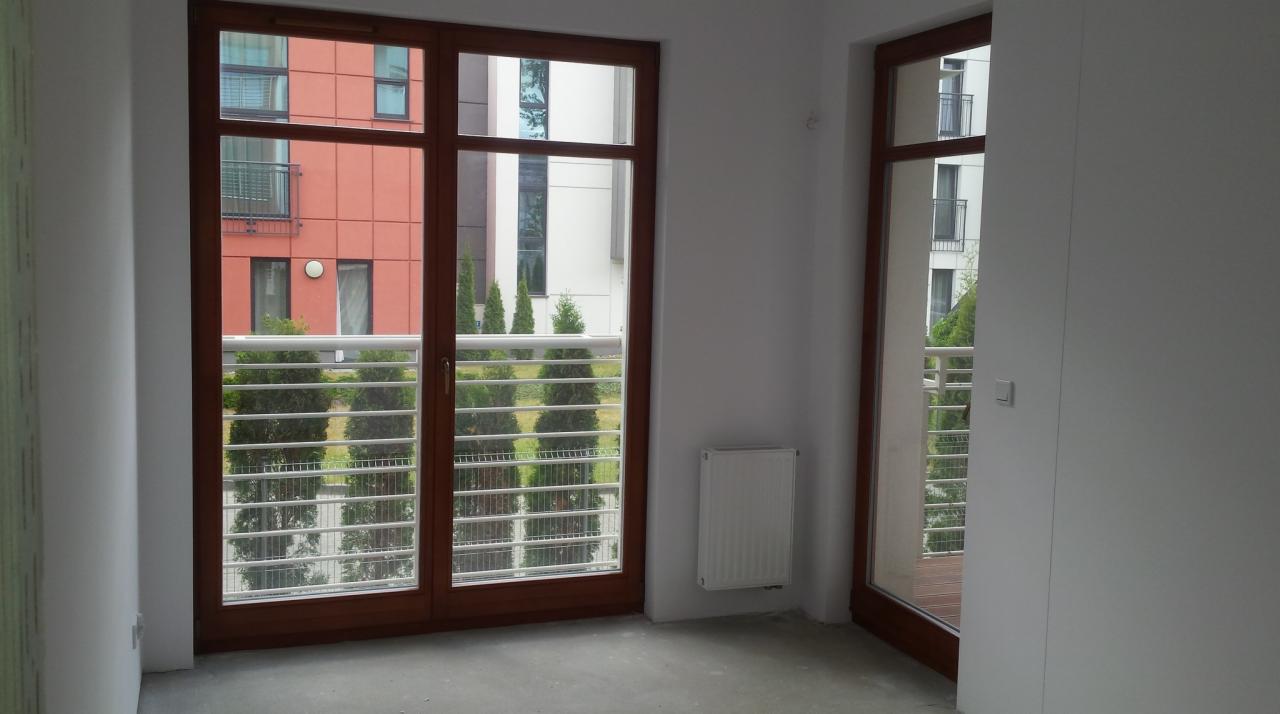 Pusty pokój z dwoma oknami balkonowymi z brązowymi ramami, widok na balkony i zieleń na zewnątrz, szare surowe podłoże, białe ściany z grzejnikiem.