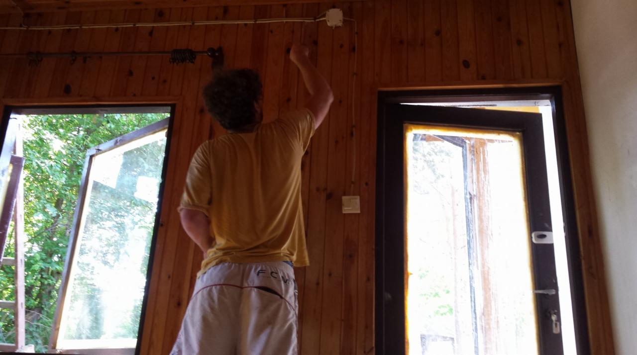 Mężczyzna w koszulce maluje drewniane ściany w pokoju z otwartym oknem i drzwiami, widoczne kable elektryczne i puszka na ścianie.