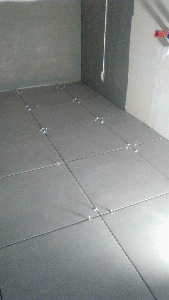 Szare płytki podłogowe w trakcie układania w rogu pomieszczenia, z widocznymi krzyżykami dystansowymi.