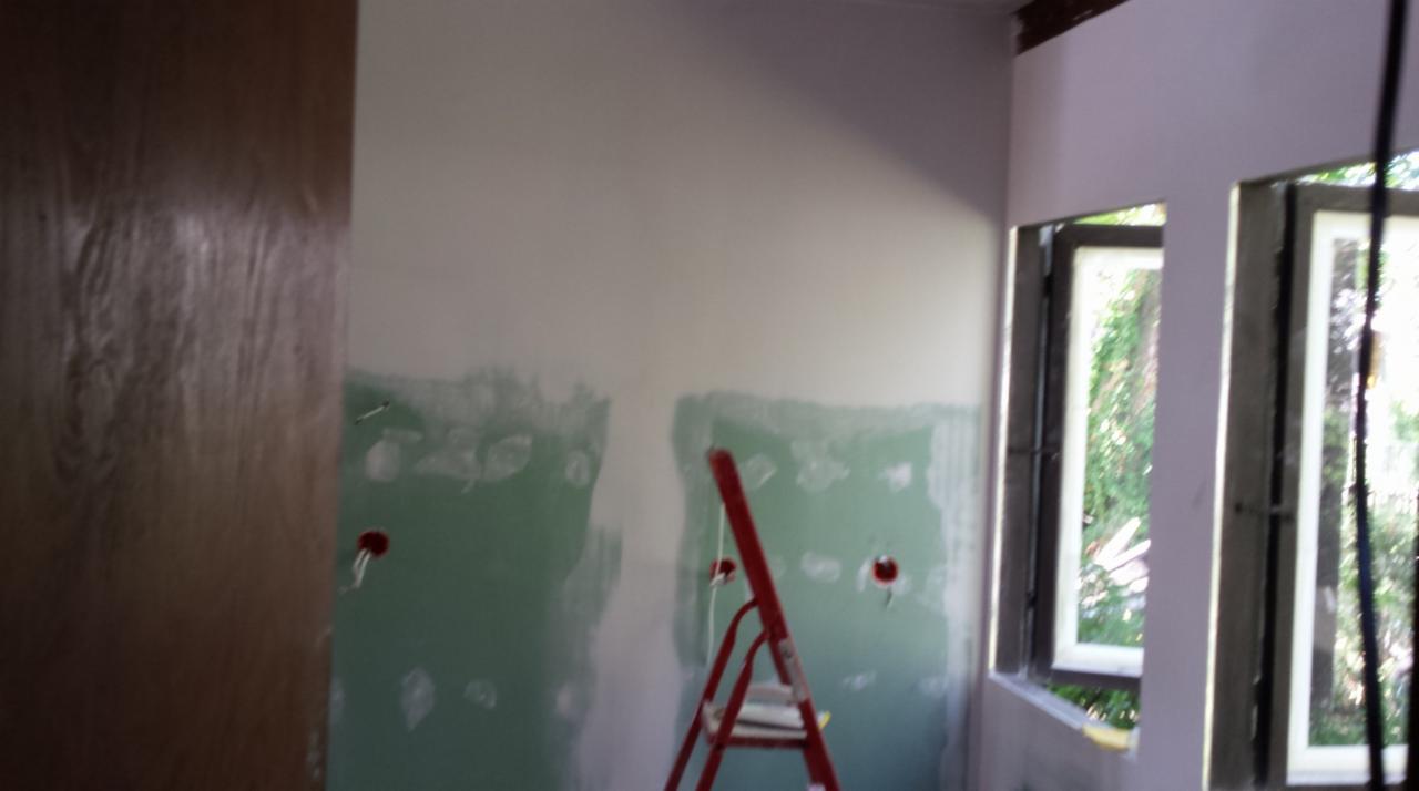 Pokój w trakcie prac wykończeniowych: ściany z widocznymi śladami szpachlowania, czerwona drabina, okna z widokiem na zieleń. Otwory po gniazdkach elektrycznych.