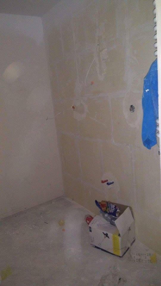Surowy stan łazienki w trakcie prac: widoczne ślady po instalacjach elektrycznych i hydraulicznych na ścianach, karton z resztkami jedzenia i butelką wody na podłodze.