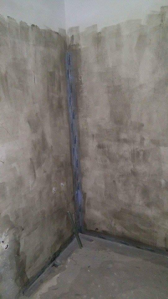 Surowe ściany w rogu pomieszczenia w trakcie przygotowania do dalszych prac wykończeniowych, widoczne listwy przypodłogowe i pionowa listwa narożna.