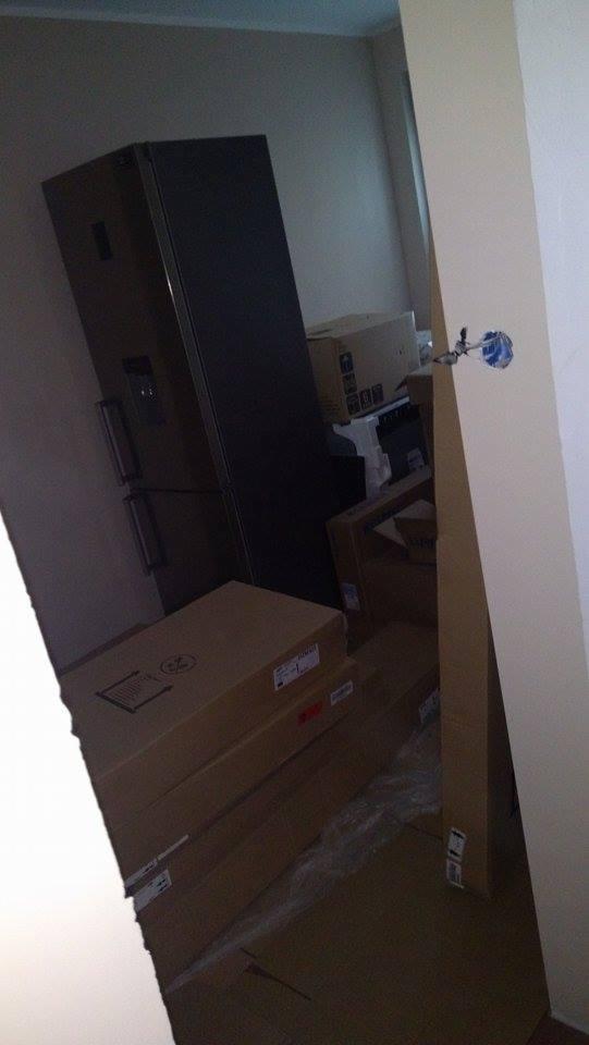 Nowa, jeszcze nie rozpakowana lodówka dwudrzwiowa w ciemnej obudowie, stoi w rogu pokoju z kartonami i przygotowaną instalacją elektryczną.