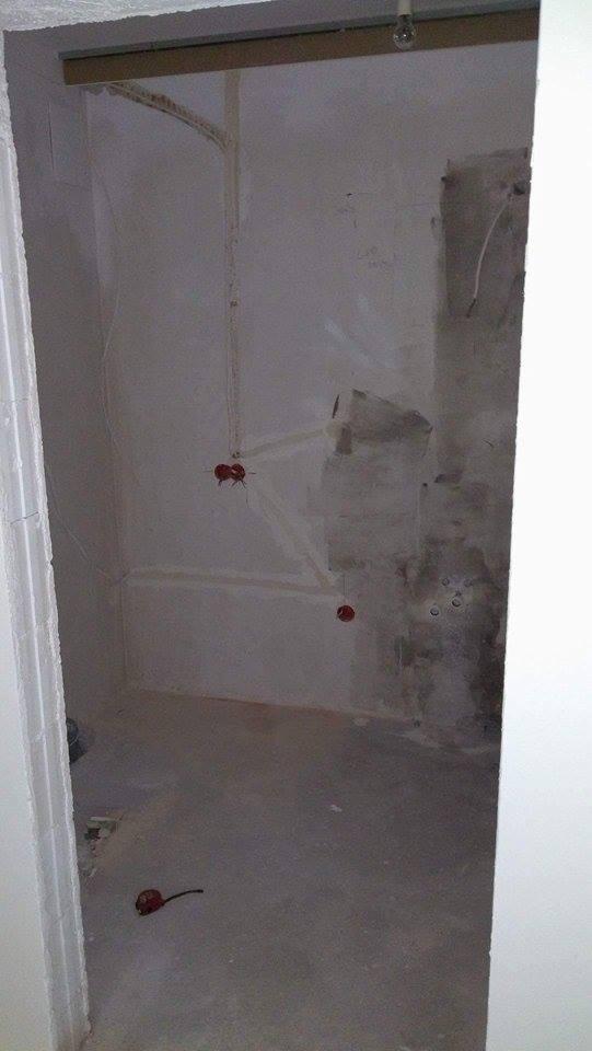 Surowe wnętrze łazienki w trakcie prac: widoczne instalacje elektryczne z puszkami, fragmenty starego tynku na ścianach i surowy beton na podłodze. Na podłodze leży czerwona miarka.