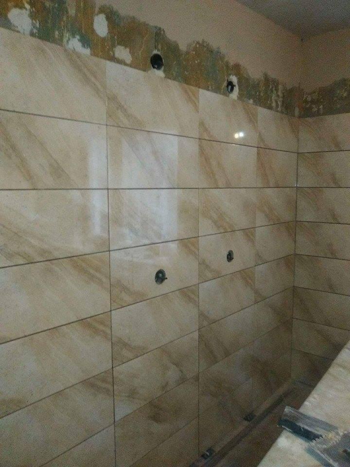 Ściana w trakcie układania beżowych, marmurkowych płytek w łazience, widoczne otwory na instalacje i niezabezpieczona górna część ściany z pozostałościami po starym wykończeniu.