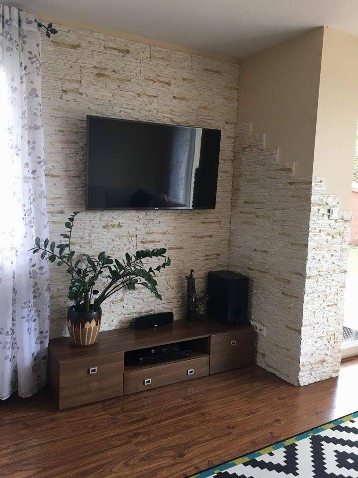 Telewizor zawieszony na ścianie wyłożonej dekoracyjną, jasną cegłą, z drewnianą szafką RTV poniżej, na której stoi roślina w ozdobnej doniczce i głośnik, na podłodze panele imitujące drewno.