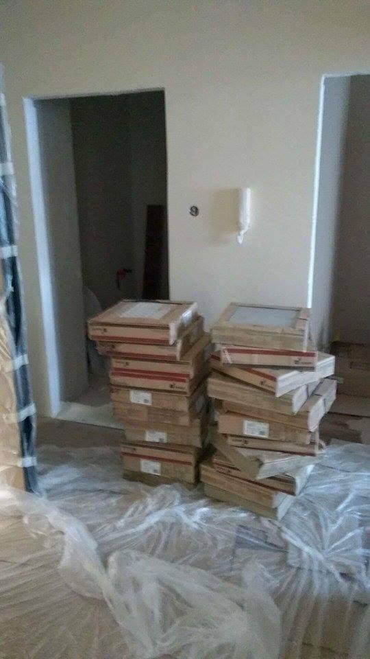 Paczki z płytkami ceramicznymi ułożone w stosy na podłodze zabezpieczonej folią malarską w trakcie prac wykończeniowych, widoczne surowe ściany i framugi drzwi.