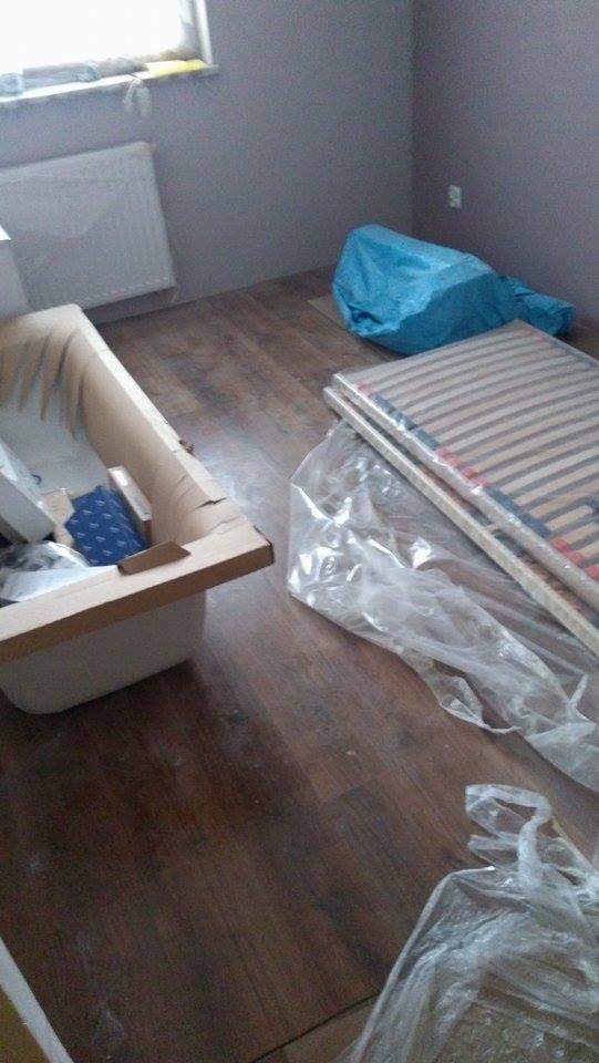 Nowa wanna w kartonie, stelaż łóżka w folii oraz niebieski worek na podłodze z paneli w pokoju z szarą ścianą i grzejnikiem pod oknem, przygotowania do remontu.