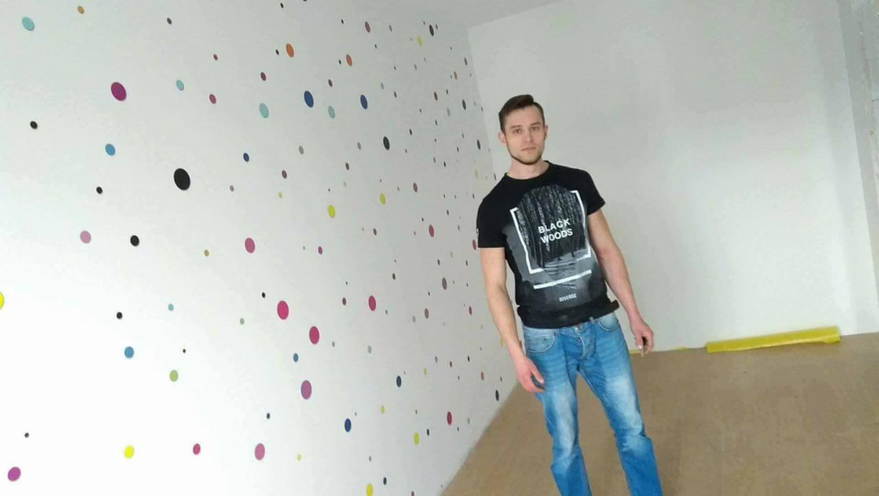 Mężczyzna w czarnej koszulce stoi na tle ściany z kolorowymi kropkami i surowej, brązowej podłogi, widoczny fragment żółtej folii ochronnej.