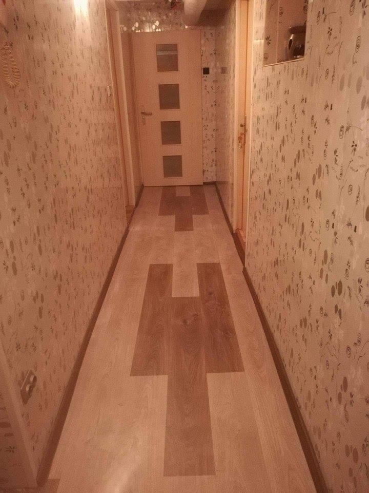 Wąski korytarz z wzorzystą tapetą w odcieniach beżu i brązu oraz panelami podłogowymi z wkomponowanym wzorem w kształcie litery H.