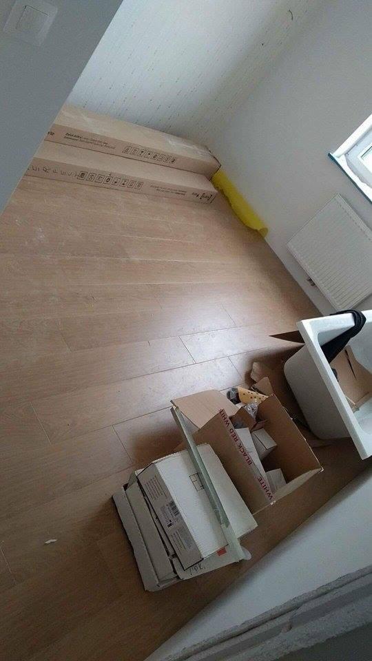 Nowa drewniana podłoga w łazience w trakcie układania, widoczne kartony z panelami, fragment umywalki i grzejnika.