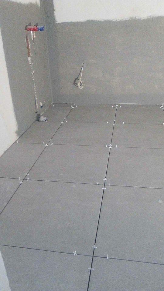 Szare płytki podłogowe w trakcie układania w łazience, widoczne krzyżyki dystansowe, surowe ściany przygotowane do wykończenia, wystające kable i rury.