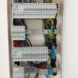 Otwarta skrzynka rozdzielcza z zainstalowanymi bezpiecznikami, przewodami w różnych kolorach i listwami zaciskowymi, widok z góry.