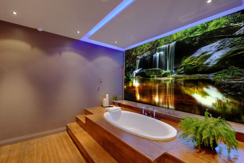 Nowoczesna łazienka z podświetleniem LED i fototapetą z motywem wodospadu, wanna na drewnianym podeście, roślina doniczkowa.