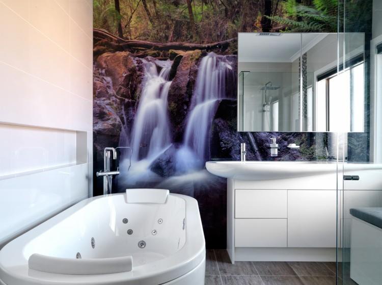 Nowoczesna łazienka z białą wanną z hydromasażem i fototapetą imitującą wodospad w lesie, odbijającą się w lustrze nad umywalką.