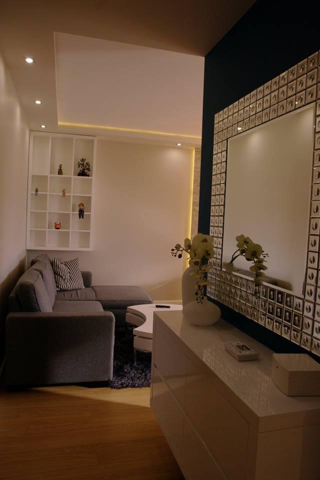 Szary narożnik z poduszką w paski, biały stolik kawowy i komoda z dekoracyjnym lustrem w ramie z kwadratowych kryształków, oświetlenie punktowe sufitowe i półka z dekoracjami.