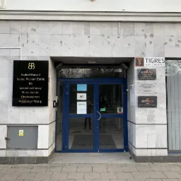 Wejście do biurowca z kilkoma tablicami informacyjnymi, w tym tablicą z nazwami marek odzieżowych i tablicami firm Tigres Nieruchomości oraz Reppo Group.