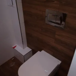 Nowoczesna toaleta podwieszana z chromowanym przyciskiem spłukującym na tle płytek imitujących drewno, białe płytki na ścianie po lewej stronie, widoczne elementy instalacji.