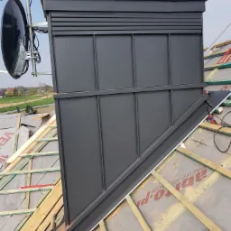 Montaż obróbki komina z blachy na dachu pokrytym membraną, widoczne elementy konstrukcji drewnianej i antena satelitarna na kominie.