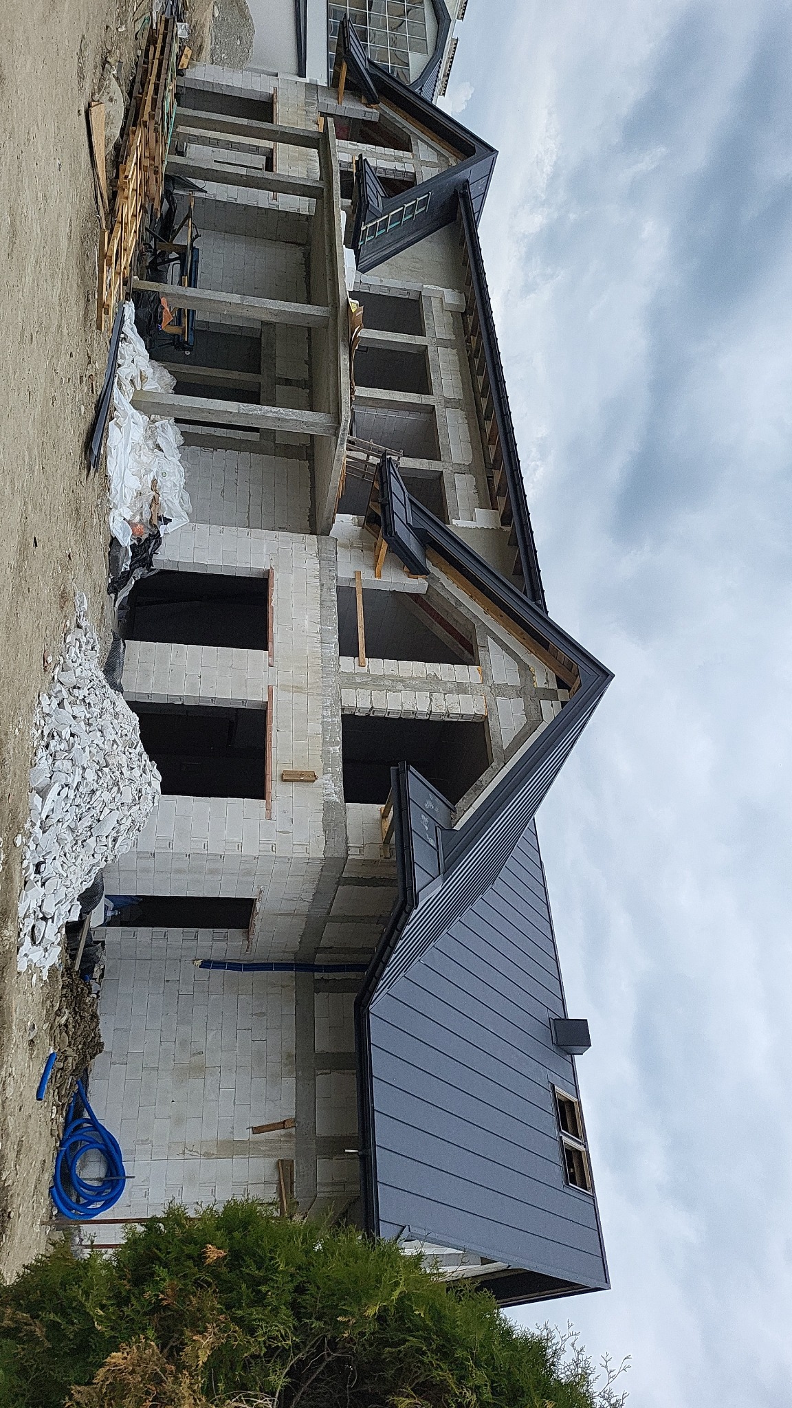 Surowy stan domu z częściowo ukończonym, grafitowym dachem z blachy na tle pochmurnego nieba. Widoczne materiały budowlane i niezabudowane okna.