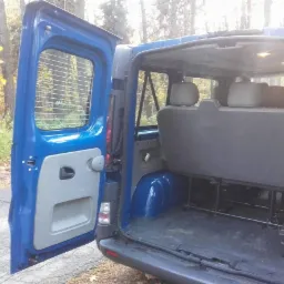 Wnętrze niebieskiego busa z otwartymi tylnymi drzwiami, widoczne trzy rzędy siedzeń i bagażnik.