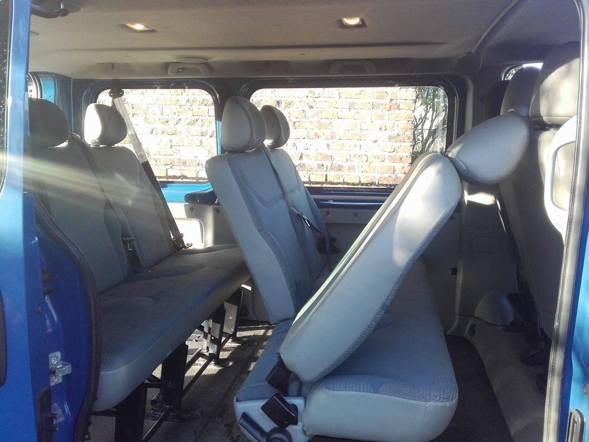 Wnętrze niebieskiego busa z szarymi siedzeniami, widok z boku przez otwarte drzwi, fotel obrócony w kierunku tyłu pojazdu, ściana z cegieł widoczna przez okno.