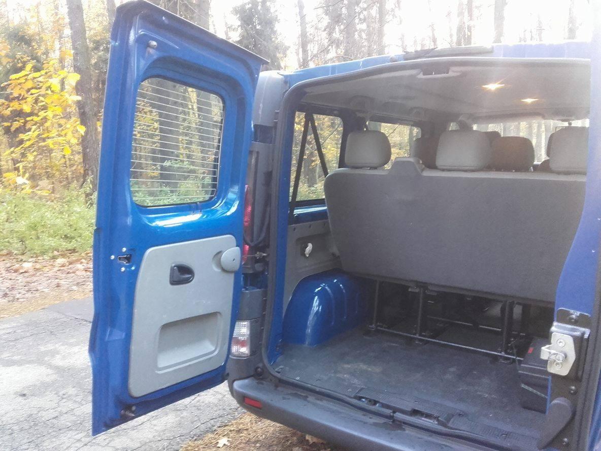 Wnętrze niebieskiego busa z otwartymi tylnymi drzwiami, widoczne trzy rzędy siedzeń i bagażnik.