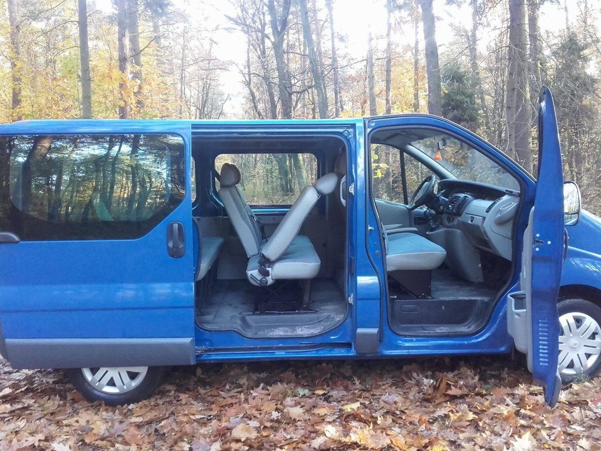 Niebieski van z otwartymi drzwiami na tle jesiennego lasu, widoczne wnętrze z szarymi fotelami.